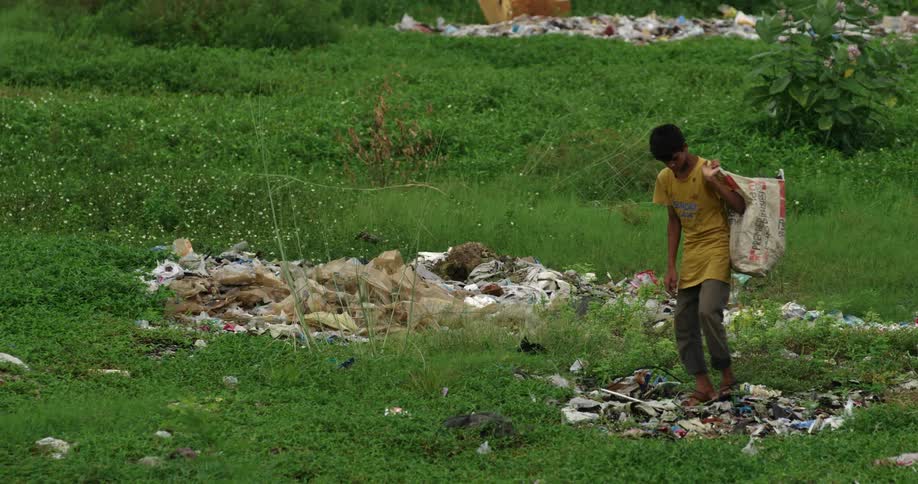 A boy searches a garbage strewn field for valuable trash to collect in his bag