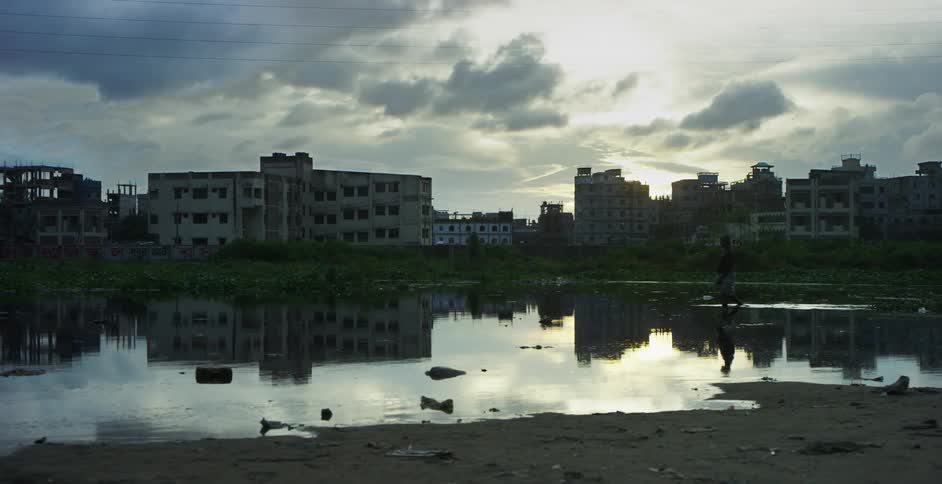 A person walks on the shallow waters of a pond near a third world country city