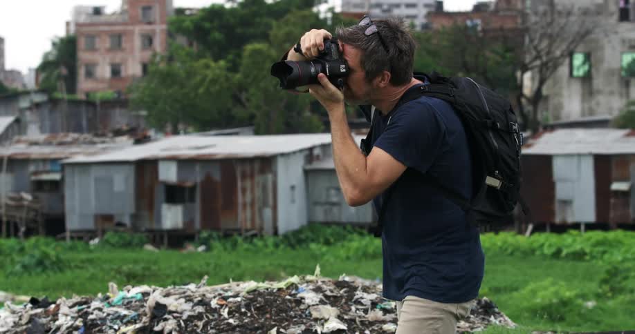 A man with a backpack a camera and glasses taking pictures with buildings and trees in the background