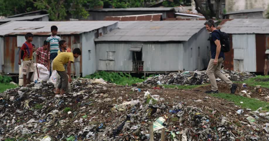A man carrying a camera walks up to a group of young boys digging through a pile of trash in a village
