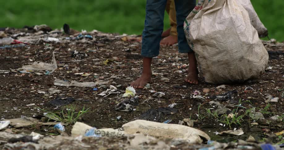 A barefoot boy collects garbage from the ground and places it in a plastic bag