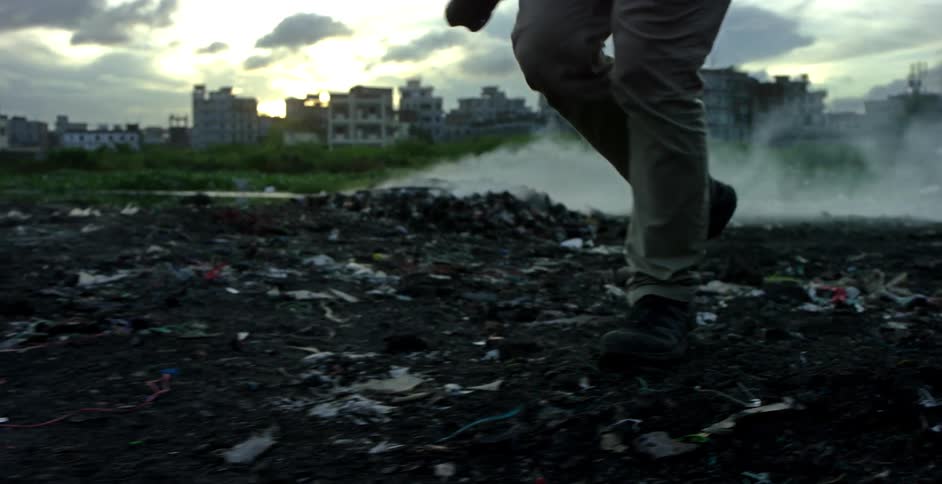 A person with a camera walking on dirt and litter with buildings in the background