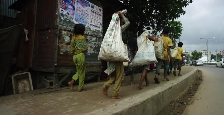 Children carrying bags in a primitive city walk by shops as cars drive past them