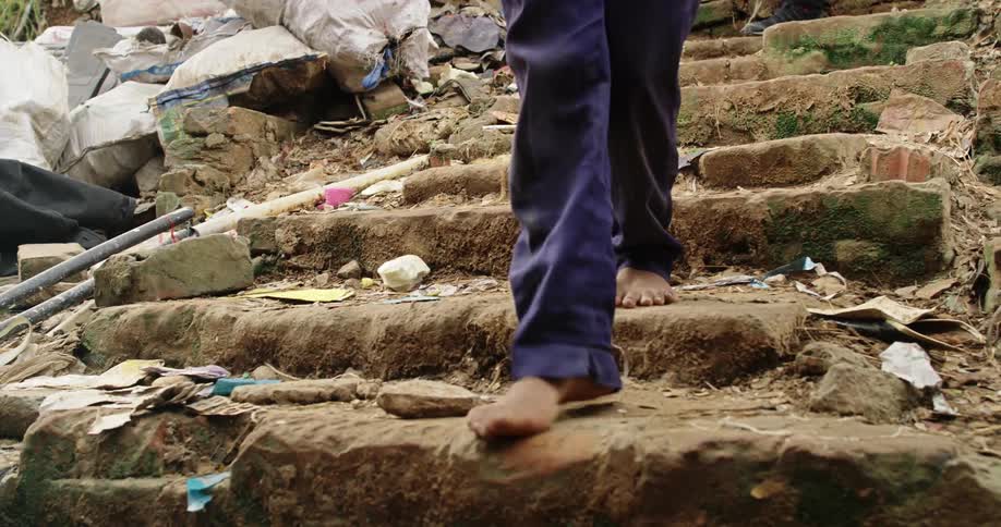A group of barefoot people carry canvas bags down an outdoor stone staircase covered in dirt moss and trash