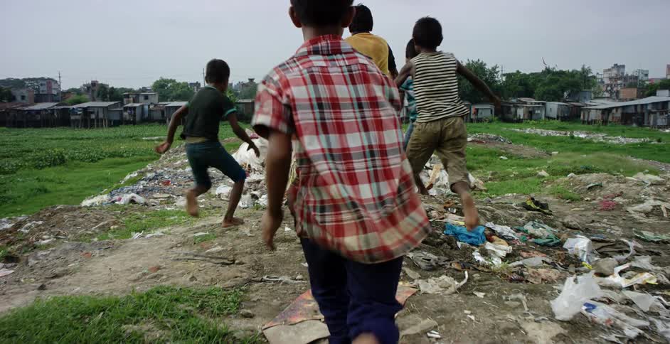 A group of boys run through a field and jump onto a heap of trash outside a slum during the day