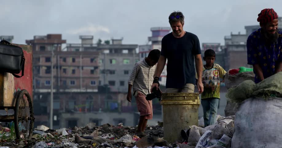 A caucasian male photographer walks over the trash covered ground at a landfill while trailed by two young children