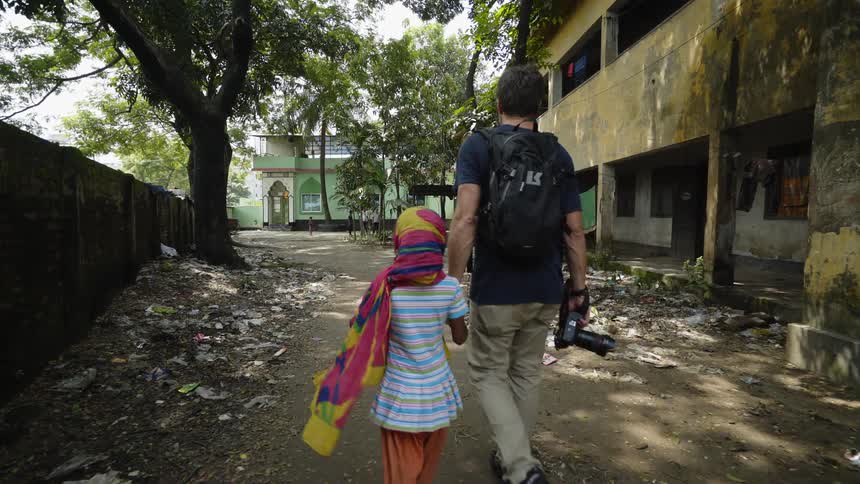 A man carrying a camera holds a young girls hands as they walk down a path past a beaten building