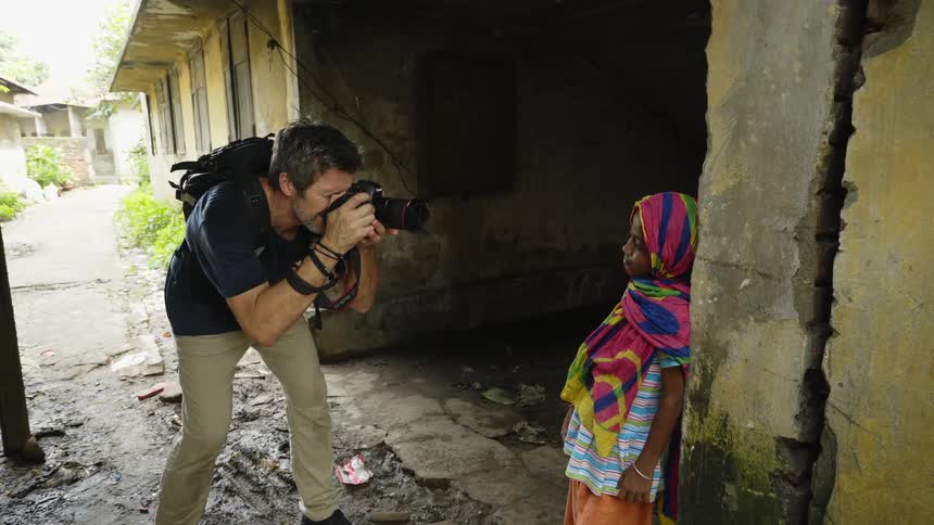 A middle aged photographer bends to take a photograph of a small girl leaning against a building