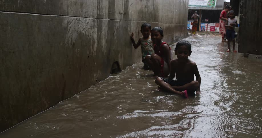 Children in a poverty stricken village play in the rain and sit down in a flooding path