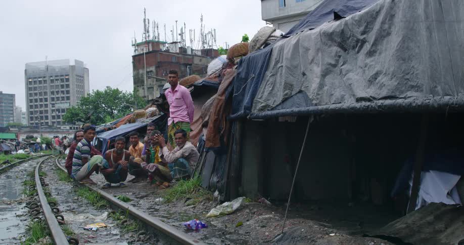A group of men sitting beside a muddy track track in the slums of a city look into the camera