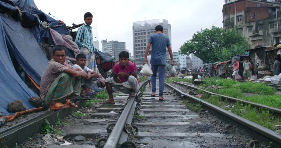 Children in a poverty stricken village sit on train tracks as a man carrying plastic bags walks past them and women by huts watch