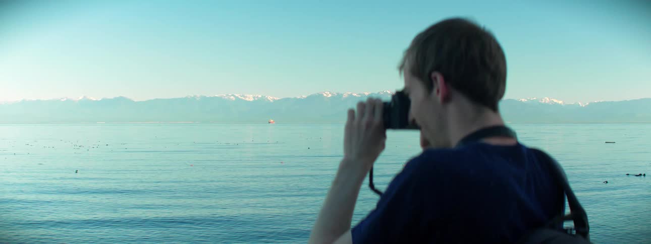 A photographer standing a lakeshore raises his camera to take a photograph of the lake and the mountains beyond it