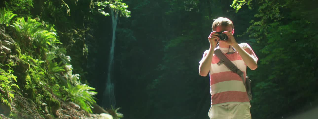 Man takes photographs by facing the camera in the forest with trees and a waterfall in the background