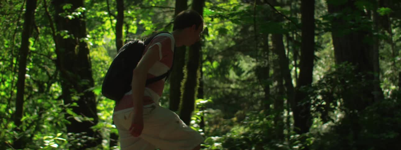Man with a t shirt and a black backpack walks in a dense forest