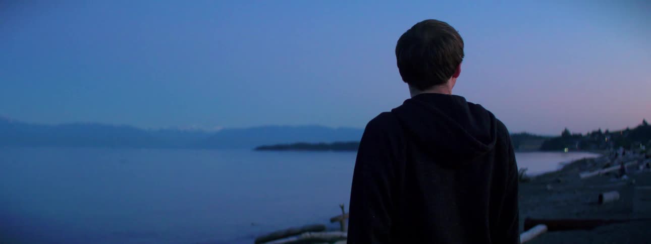 A male stands slightly above a coastline that is met by serene waters