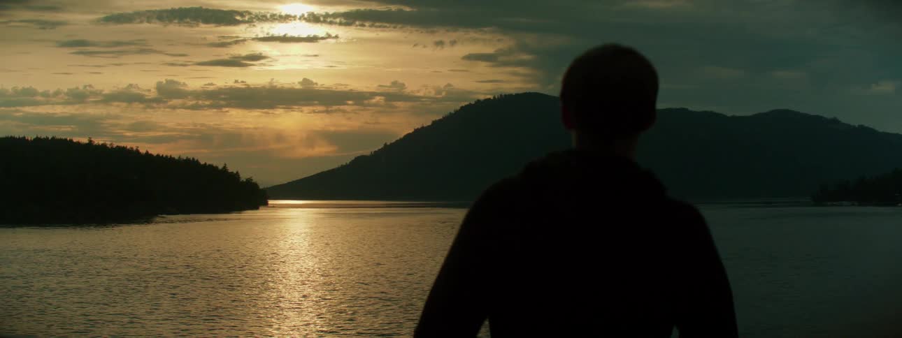 Mans silhouette looks over water reflecting light toward mist covered forested mountains