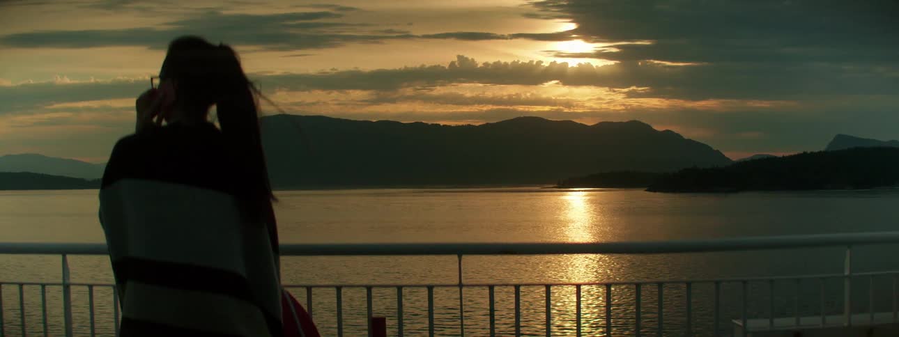 Woman near the railing of a boat looks at the mountains at sunset
