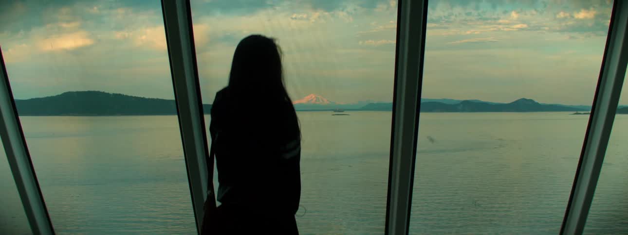 A woman stands in front of a bank of windows and looks out over a body of water
