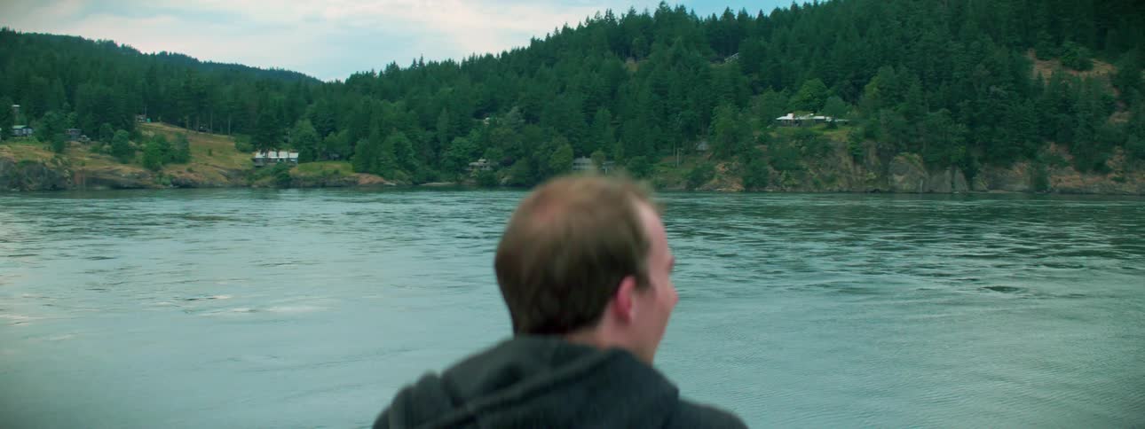 A man in a hoodie looking at a lake with trees and the sky in the background