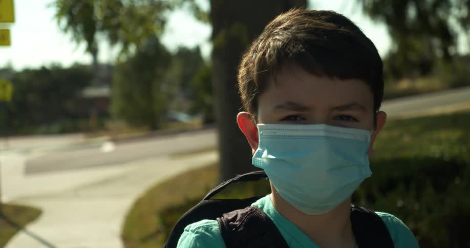 Boy wearing backpack and face mask stands on sidewalk near school
