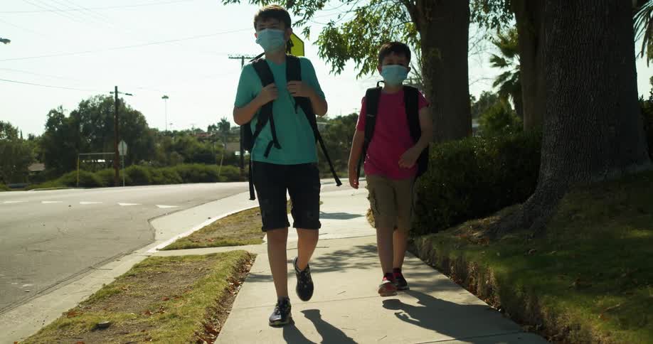 Two boys with backpack and blue masks walks on the sidewalk next to the road