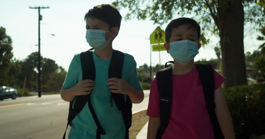 Two boys with backpacks and blue masks walk on the sidewalk next to the road