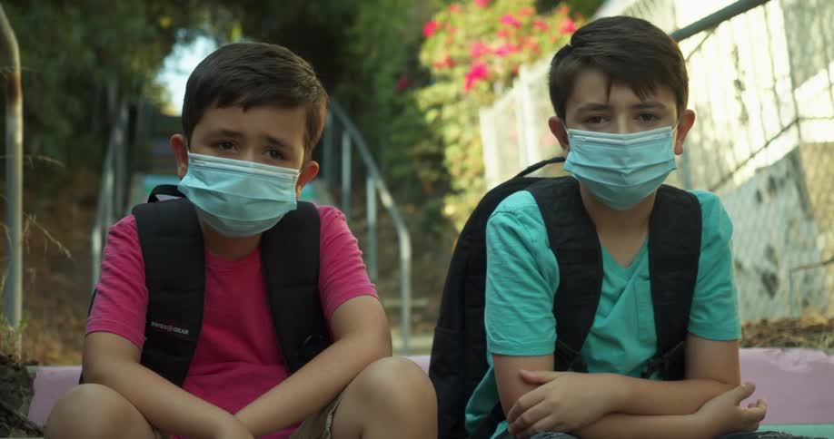 Two boys with blue masks sit down and face the camera with eye contact in a park with colorful stairs in the background