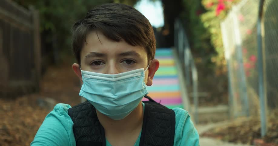 Boy with a blue mask faces the camera with eye contact in a park with colorful stairs in the background