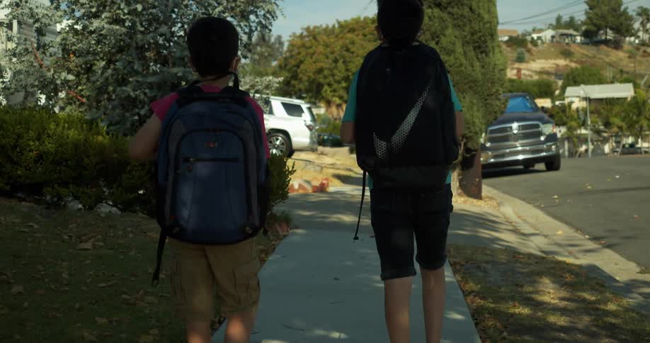 Two young boys wearing backpacks walk on the sidewalk of the neighborhood