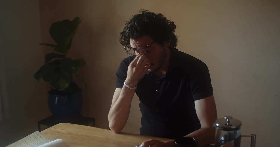 A man sits at a table with a book and wipes his eyes out of frustration or tiredness