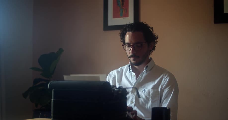 Man with glasses a beard and a white shirt sits at a desk and uses an old typewriter