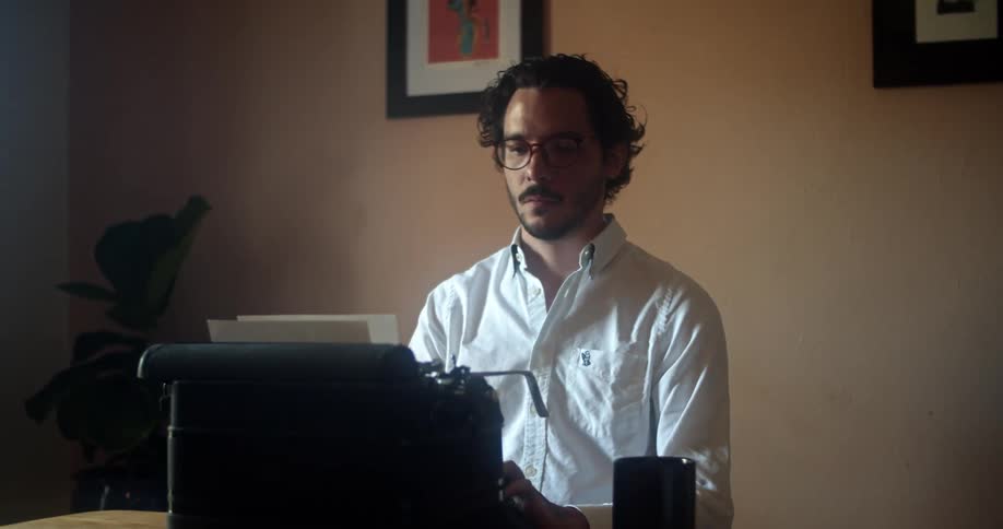 A man with glasses sitting on a chair in front of a table with a typewriter