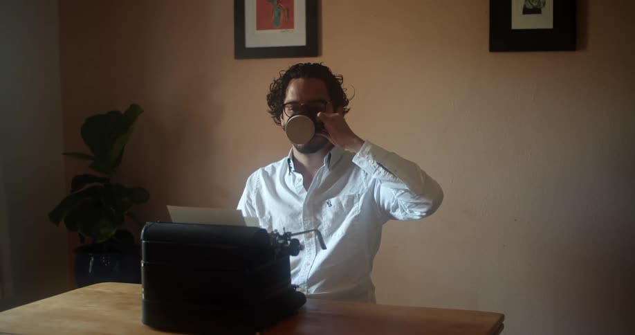 Man with glasses beard and a white shirt sits at a table and uses an old black typewriter while drinking a cup fo coffee