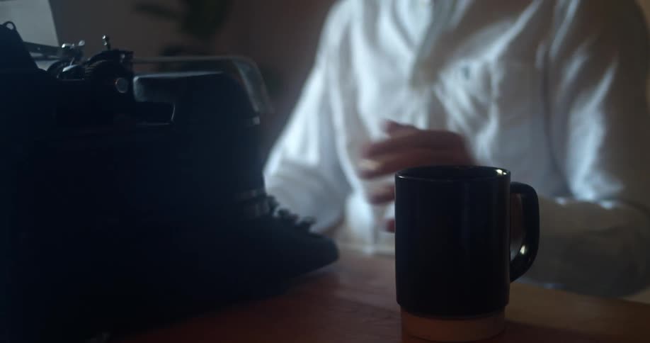 Man with a white shirt sits at a desk and uses a black old typewriter while drinking a cup of coffee