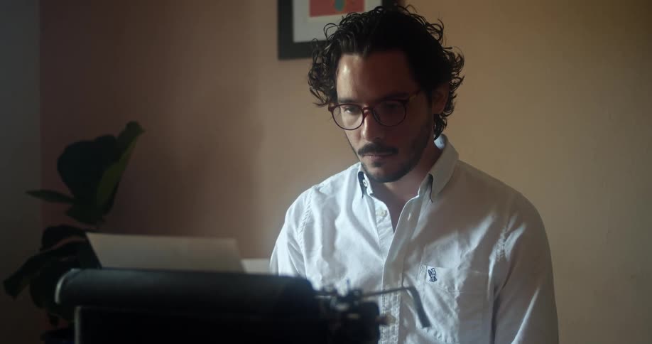 Young man wearing a white shirt and glasses sits down in front of a typewriter in a room