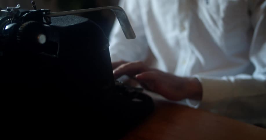 Man with a white shirt sits at a desk and uses a black old typewriter