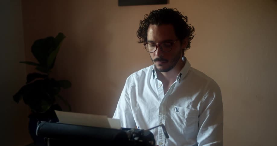 Man with glasses a beard and a white shirt uses an old black typewriter in a room