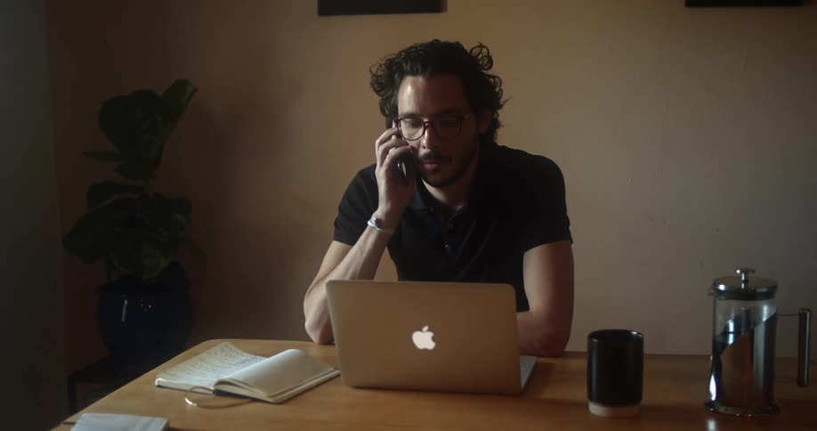 Man with glasses a beard and a black polo shirt sits at a table and makes a phone call with a smartphone in a room