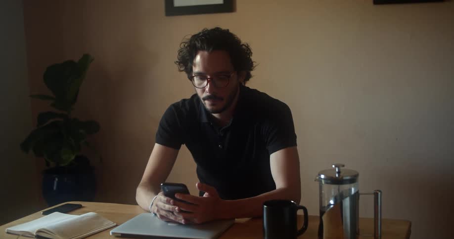Man with glasses a beard and a black polo shirt closes his laptop and uses his smartphone in a room