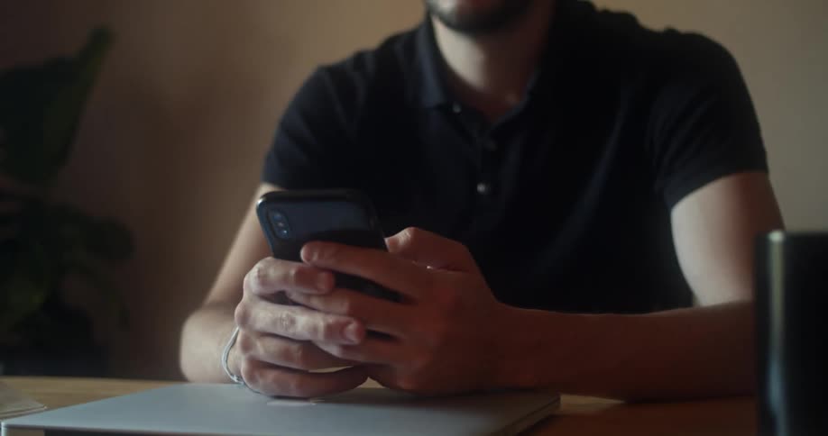 Man with glasses a beard and a black polo shirt sits at a table and uses a smartphone smilingly