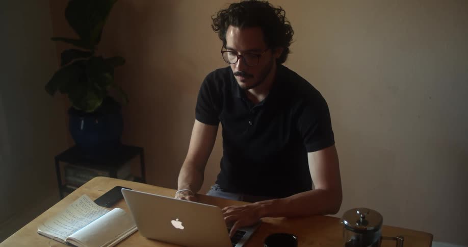 Man with a black polo shirt sits at a table and uses a grey macbook laptop in a room
