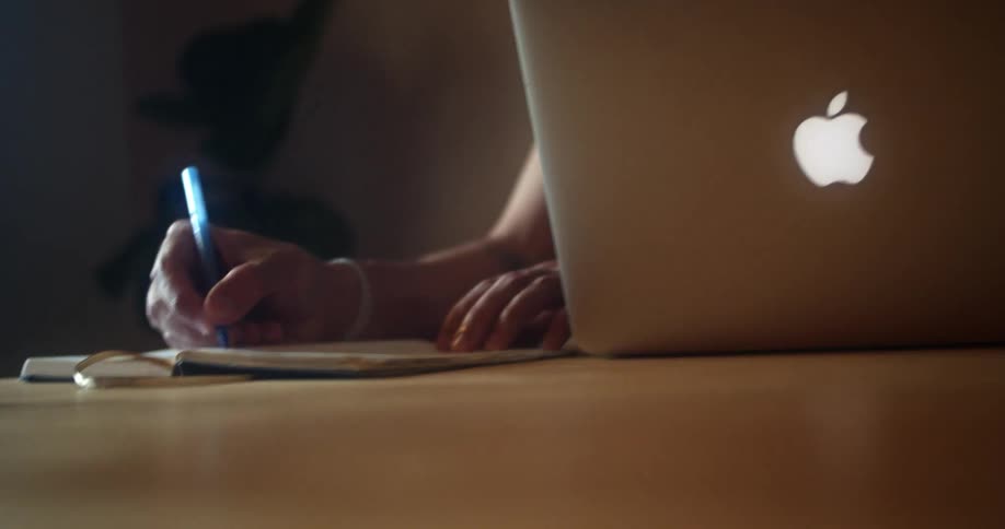 Man with glasses a beard and a black polo shirt sitting at a table writes in a notebook and uses a laptop in a room