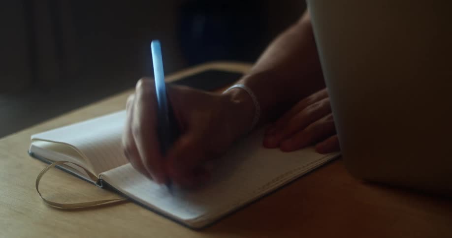 Mans hand writes in a notebook between a smartphone and a grey laptop in a room