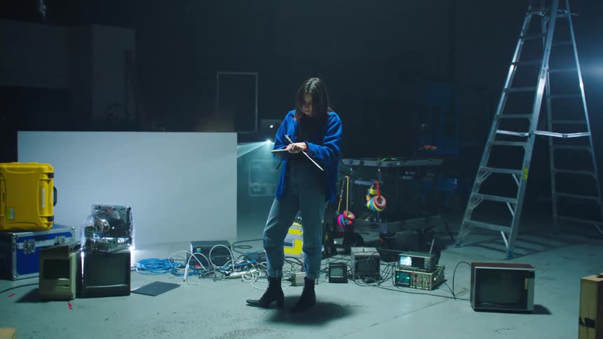 A woman on a film set writes on a clipboard while being surrounded by electronics and props