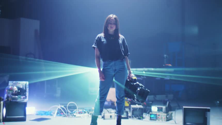 A woman stands on a stage with film equipment and televisions and looks around