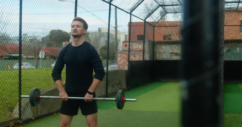 A man lifts a barbell in a caged off area in a city during the day