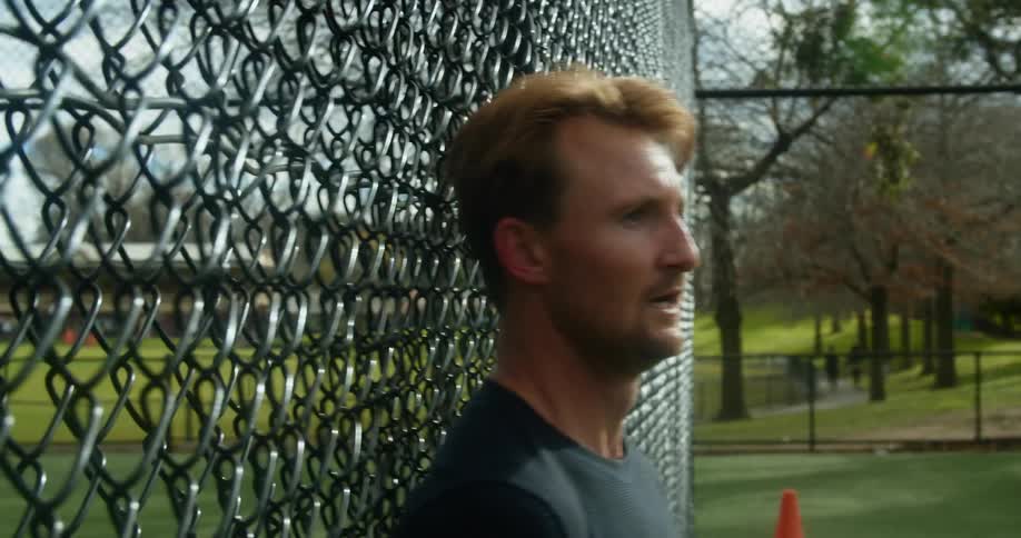 A man in a public park falls against a fence and heavily breathes during exercise while people stroll along a path in the background