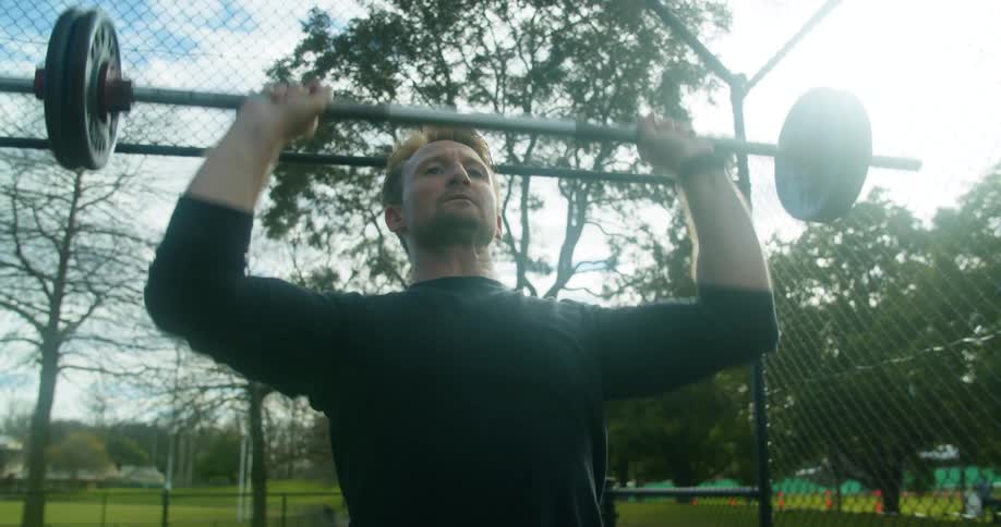 A man lifts a barbell up and down at an outside exercise space