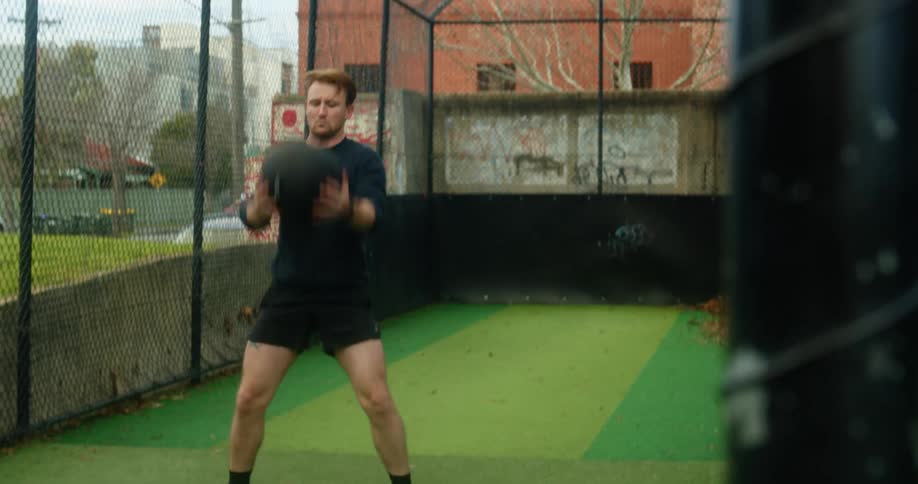 A man throws a weighted ball behind him in his exercise routine in an outside cage