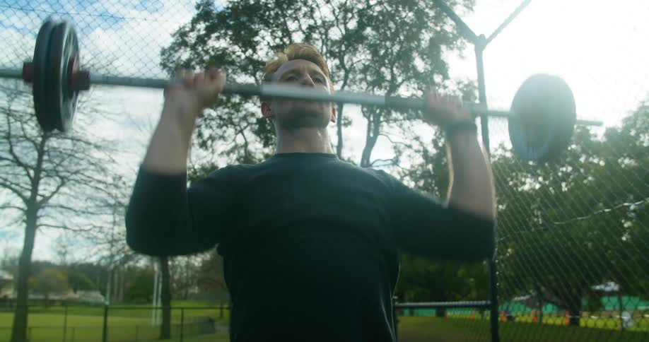 A man in a public park lifts weights as kids play in the background